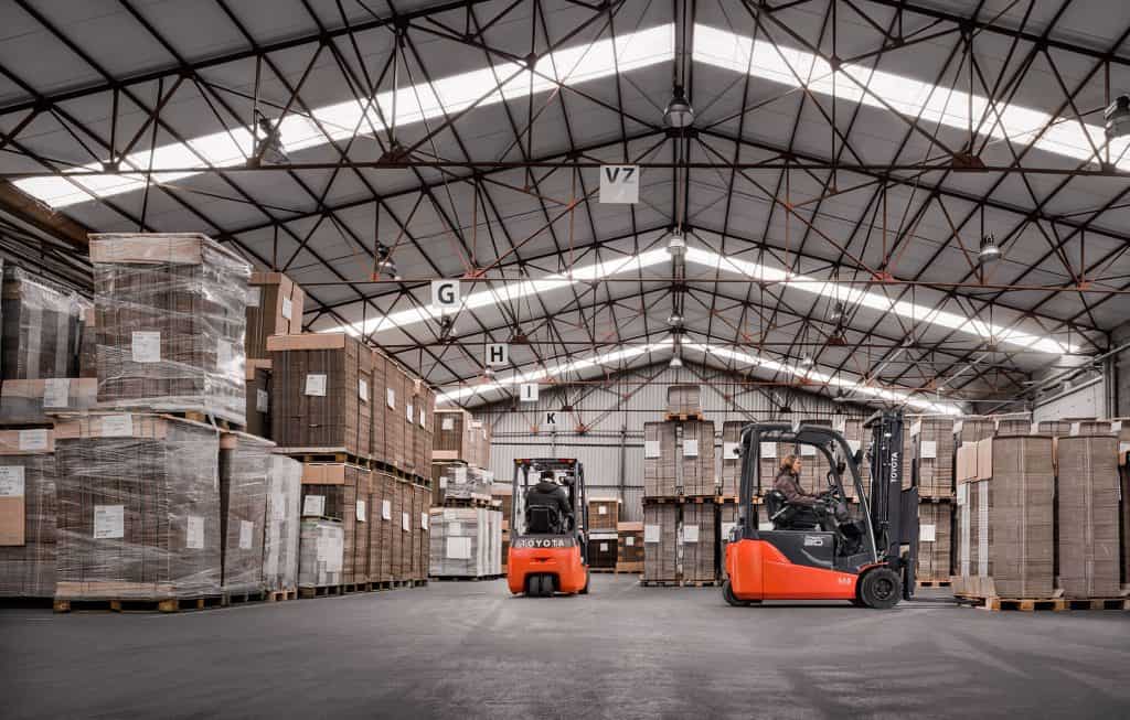 Forklift trucks operating in a large warehouse with stacked pallets and high ceilings.